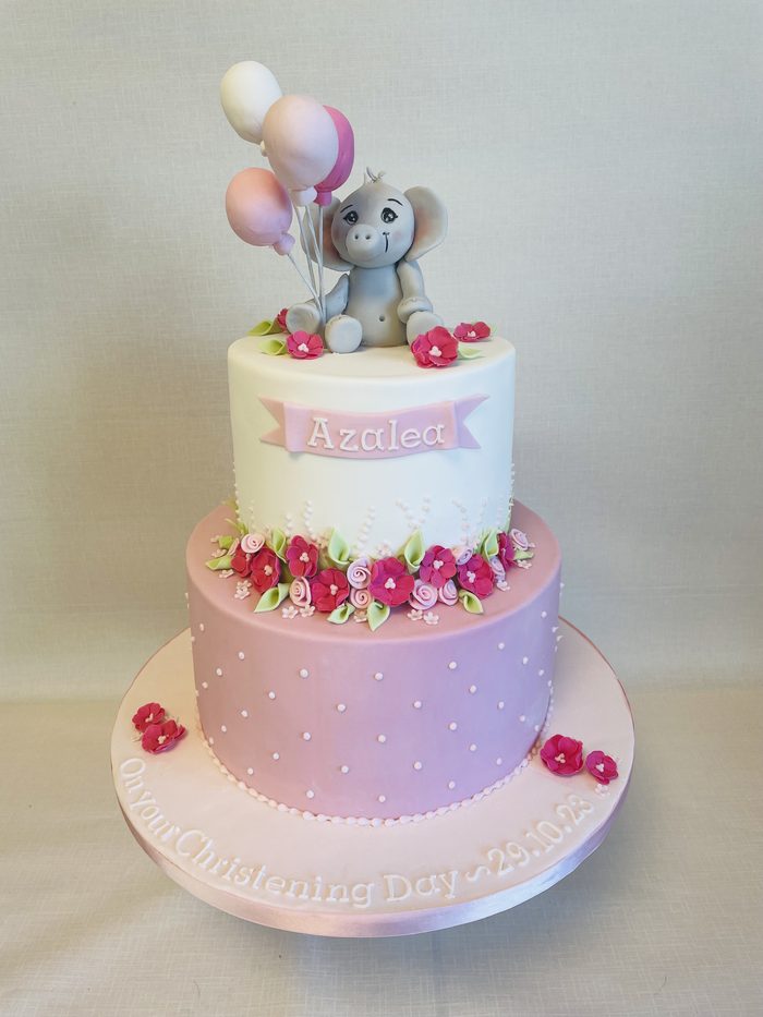Two tier pink ombre christening cake with sugar flowers and an elephant topper holding balloons
