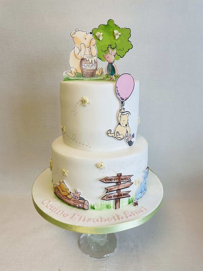 Two tier christening cake with classic Winnie the Pooh characters, a signpost and a balloon